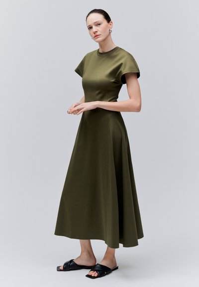 Femme aux cheveux foncés attachés en chignon bas, portant une robe midi à manches courtes couleur vert olive et des sandales noires à enfiler, se tenant devant un fond clair uni.