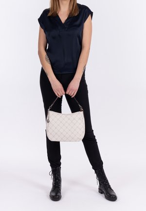 Femme portant un pantalon noir, des bottes noires et un chemisier bleu marine à manches courtes, tenant un sac à main matelassé blanc devant un fond uni.
