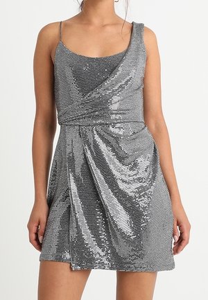 Vestido mini de lentejuelas plateadas con una tira fina y una tira caída en el hombro, cintura drapeada, llevado por una persona de pie contra un fondo liso.