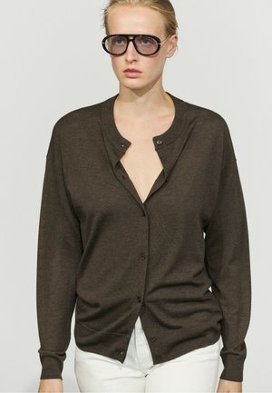 Femme portant de grosses lunettes noires et un cardigan marron foncé boutonné sur un pantalon blanc, sur un fond clair uni.