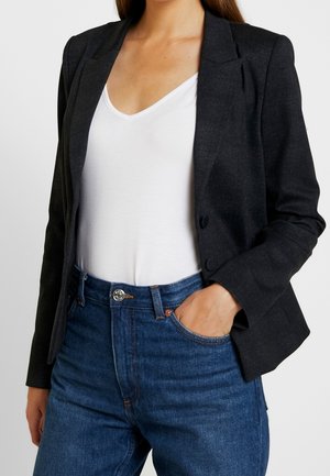 Kvinde iført mørkegrå blazer over en hvid V-hals t-shirt, blå jeans, med venstre hånd i jeanslommerne, overkroppen synlig, hoved ikke vist.