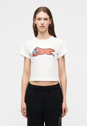 Γυναίκα με καστανά ίσια μαλλιά φοράει λευκό κοντό t-shirt με κόκκινη καρτουνίστικη αλεπού και μαύρο παντελόνι σε απλό γκρι φόντο.