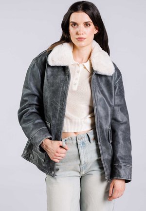 Veste en cuir gris avec un col en fausse fourrure blanche duveteux, fermeture éclair à l'avant, deux poches latérales et denim légèrement usé. Top court beige clair.