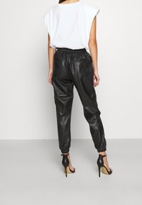 Kvinna klädd i en ärmlös vit topp, svarta högmidjade läderbyxor och svarta högklackade sandaler, stående mot en enkel bakgrund.