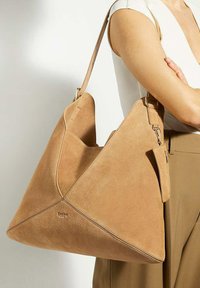 Tan suede handbag with a triangular design, flat base, long adjustable strap, and a small attached zip pouch. Subtle logo detail visible.