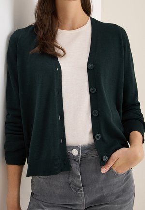 Frau trägt einen dunkelgrünen, geknöpften Cardigan über einem weißen Hemd, mit der Hand in der Tasche grauer Jeans, gezeigt von den Schultern bis zur Hüfte.