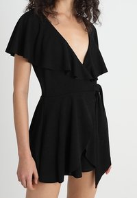 Mujer vistiendo un vestido negro envolvente con mangas con volantes y un escote en V profundo, que muestra parte de su muslo medio y brazos.