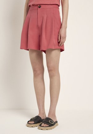 Coral pink shorts with a high waist, pleated front, and side pockets. Worn with black slide sandals featuring a gold accent buckle.