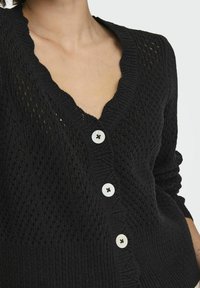 Black knit cardigan featuring a scalloped V-neckline, textured pattern, and three large white buttons. Accents include ribbed cuffs.