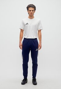 Νέος άνδρας στέκεται μπροστά φοράει λευκό αθλητικό πουκάμισο, μπλε σκούρο παντελόνι φόρμας με λευκές ρίγες και μαύρα αθλητικά παπούτσια σε απλό λευκό φόντο.