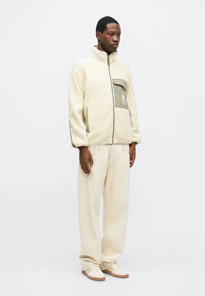 Fleece zip-up jakke i krem med en kontrasterende olivengrønn lomme, paret med beige vide bukser og lyse joggesko.