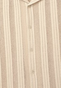 Lys beige skjorte med knaplukning, der har en vævet tekstur med lodrette striber og små vekslende åbninger. Inkluderer to knapper.