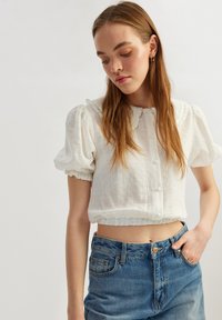 Hvid cropped bluse med korte puffærmer og flæsekrave, der har en knaplukning foran og tekstureret stof. Sammen med blå denimjeans.
