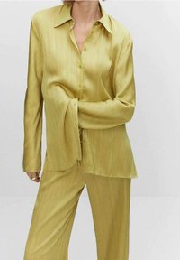 Chemise boutonnière jaune à manches longues avec une texture rayée subtile, portée avec un pantalon assorti. Coupe loose, col et détails minimaux.