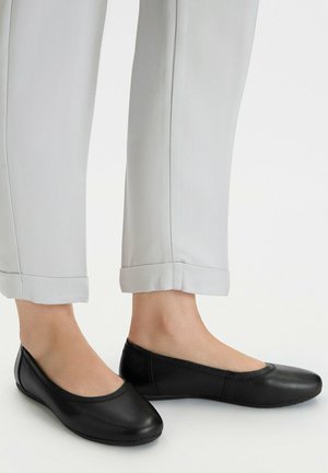 Zapatos planos de ballet de cuero negro usados con pantalones gris claro con puños, que muestran los tobillos y la textura suave del zapato sobre un fondo blanco liso.