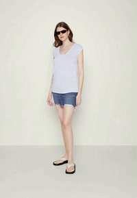 T-shirt cinza-claro em formato de V, feito de tecido suave, combinado com uma saia mini de ganga desgastada. O modelo usa óculos de sol pretos e chinelos.