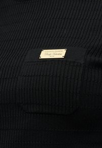 Haut noir côtelé avec un tissu texturé, une petite poche poitrine et une plaque logo de marque dorée fixée à l'avant.