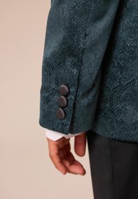Teal strukturerad blazermedel med tre matta svarta knappar, kontrasterande vit manschettdetalj, som visar upp en skräddarsydd passform och premiumtyg.