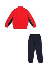 Giacca rossa con zip e strisce laterali blu, abbinata a pantaloni della tuta blu. Entrambi gli articoli sono realizzati in morbido tessuto con polsini e vita elasticizzati.