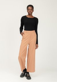 Schwarzes geripptes Langarmoberteil kombiniert mit hellbraunen weiten Hosen. Das Modell trägt schwarze Plateauschuhe mit goldenen Kettenakzenten.