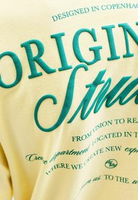 Tissu jaune avec texte en relief bleu sarcelle indiquant « ORIGINAL Studio » et texte plus petit en écriture cursive et imprimée mentionnant Copenhague et le département créatif.