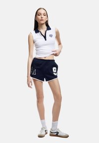 Biała bezrękawnikowa koszulka polo z granatowym kołnierzykiem, w połączeniu z granatowymi szortami z białymi wykończeniami i numerami. Model nosi białe skarpetki i tenisówki.
