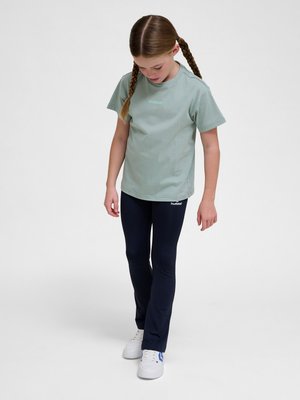 Mädchen mit geflochtenem Haar, das ein hellgrünes T-Shirt und marineblaue Leggings trägt, schaut nach unten mit entspannten Armen an den Seiten.