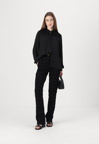 Zwart overhemd met textuur op de schouders, gecombineerd met zwarte, gestreepte broek met geometrische accenten. Model houdt een kleine zwarte handtas vast.