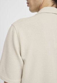 Polo de color beige con canalé y cuello corto. La tela texturizada presenta rayas verticales, realzando un corte relajado y mangas cortas.