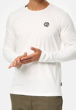 Homme portant une chemise blanche à manches longues avec un petit logo noir sur la poitrine et un pantalon vert olive, mains jointes devant.