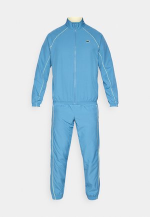 Tuta azzurro chiaro in materiale sintetico, con giacca con zip e collo alto e pantaloni coordinati con polsini elastici.