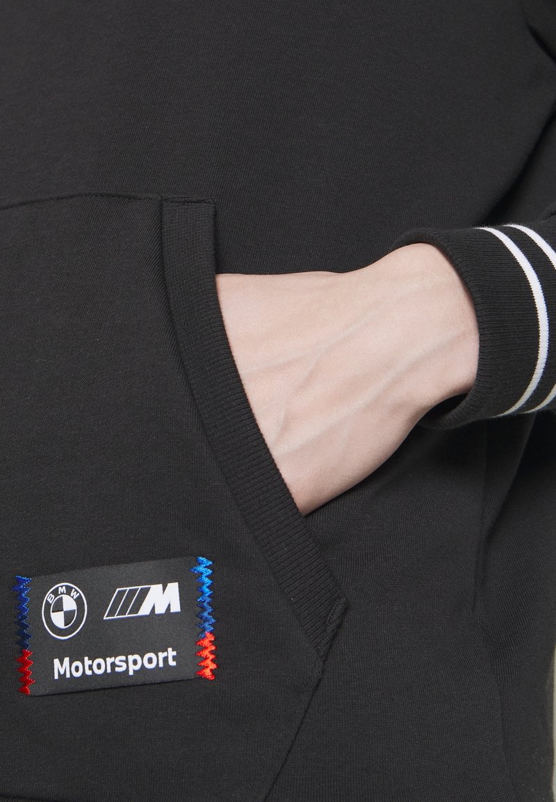 Mano nella tasca di una felpa nera BMW Motorsport con il logo a patch ricamato in blu, bianco e rosso.