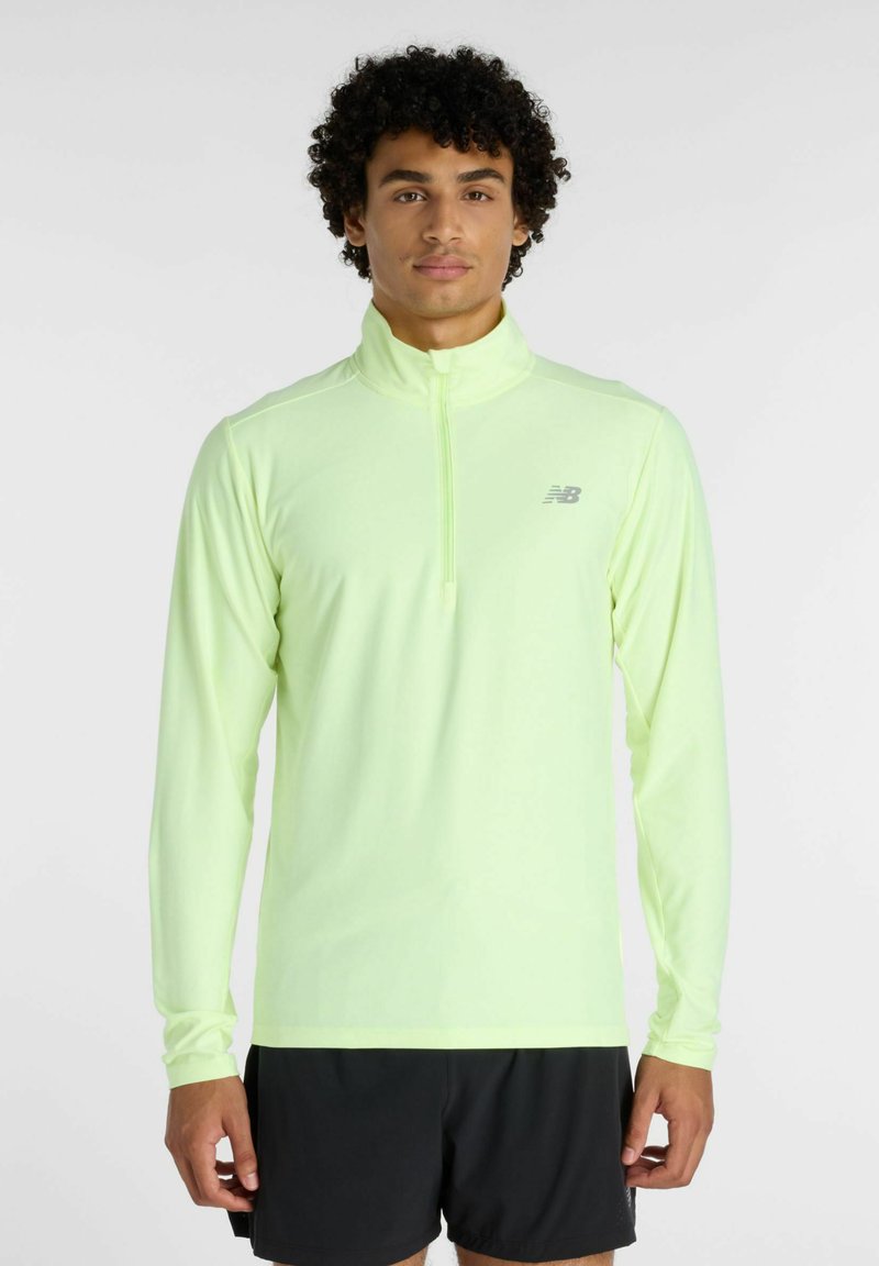 Homme aux cheveux bouclés portant un haut de sport à manches longues vert clair avec une fermeture éclair au quart et un short noir, debout devant un fond uni.