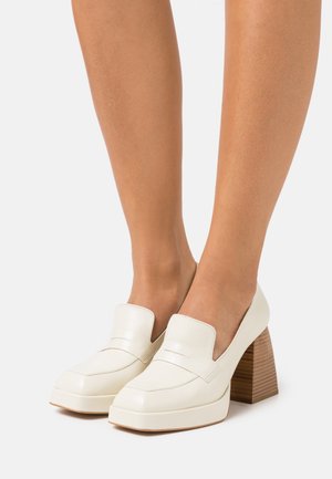 Mocassins en cuir blanc avec un bout carré, présentant un talon en bois chunky et un détail de sangle à l'avant. Le design est élégant et minimaliste.