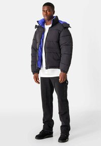 Zwarte gewatteerde jas met een blauwe binnenkant en hoge kraag. Voorzien van elastische manchetten en zijzakken. Draagt met een wit shirt en zwarte broek.