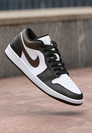 Baskets basses noires, blanches et brunes avec lacets et logo Nike, présentées en plein vol au-dessus d'une surface en pierre et en béton.