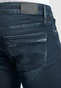 Jeans en denim bleu foncé présentant une texture lisse, un patch en cuir à la taille et plusieurs poches avec des détails de surpiqûres discrets.