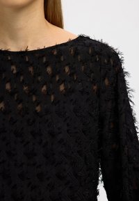 Svart texturerad topp med genomskinlig design, lager av fransiga detaljer, båtringning och långa ärmar.