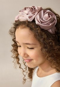 Un bandeau en satin rose clair avec deux grandes roses en tissu décoratives aux pétales enroulés, posé sur des cheveux bouclés et un fond neutre.