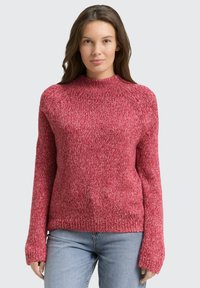 Frau trägt einen dicken, rosa melierten Strickpullover mit langen Ärmeln und hellblauen Jeans, steht vor einem schlichten hellgrauen Hintergrund.