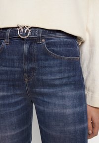 Jeans de mezclilla azul oscuro con un sutil patrón desgastado, con costuras doradas, cinco bolsillos y un cinturón con anillo decorativo de metal.