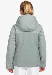 Ljust grön isolerad jacka med huva, kontrasterande övre del och elastiska manschetter. Har en liten logotyp på den nedre fållen och inga synliga mönster.