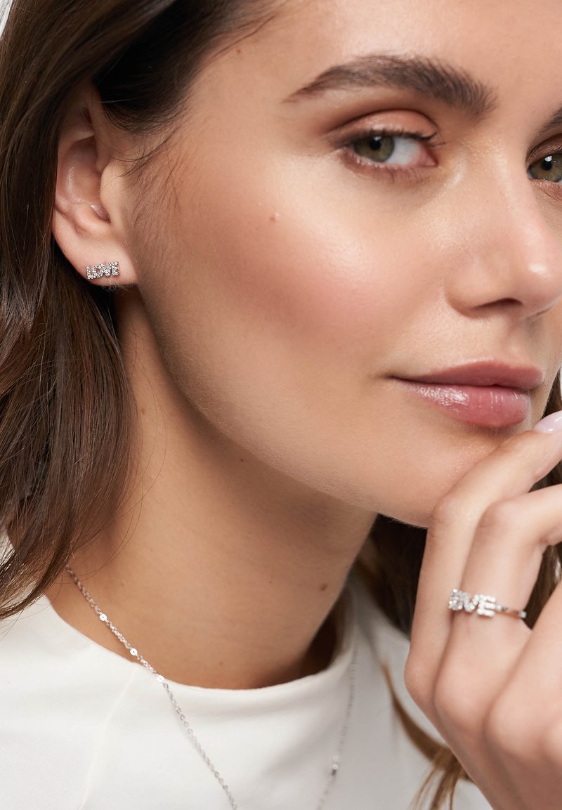 Silberner Schmuck mit "LOVE" aus Strass, getragen als Ohrstecker und Ring, mit einer glatten, polierten Oberfläche vor einem hellen Hintergrund.