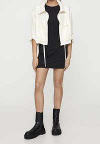 Biała kurtka bomberka ze sznurkami, noszona na czarnej dopasowanej sukience i czarnych botkach na obcasie. Gładka tkanina i czyste linie na całej powierzchni.