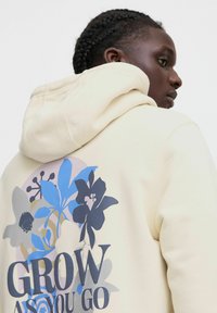Sweat à capuche crème avec un grand motif floral en bleu et noir, comportant le texte "GROW AS YOU GO" en bleu foncé. Texture de tissu douce.