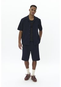 Camicia a maniche corte navy con bottoni e tessuto testurizzato, abbinata a pantaloni corti coordinati. Gli accessori includono calzini color crema e scarpe marroni.