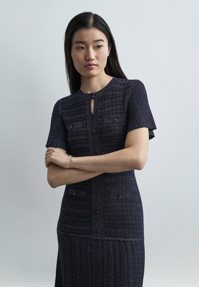 Femme aux longs cheveux foncés portant une robe courte texturée bleu marine avec boutons, debout les bras croisés devant un fond clair uni.