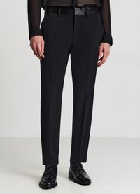 Schwarze, maßgeschneiderte Hose mit schmaler Passform, glatter Textur und Bügelfalte vorne. Kombiniert mit glänzenden schwarzen Lederschuhen.