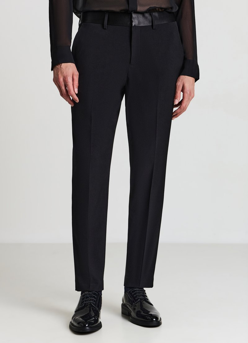 Schwarze, maßgeschneiderte Hose mit schmaler Passform, glatter Textur und Bügelfalte vorne. Kombiniert mit glänzenden schwarzen Lederschuhen.