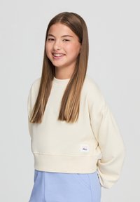 Felpa avorio tagliata con spalle basse, polsini a coste e un'etichetta rosa in basso a sinistra. Indossata con pantaloni azzurri chiari.